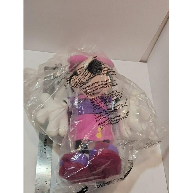 May include: A pink and purple Minnie Mouse plush toy in a clear plastic bag. The toy is wearing a pink hat, a purple shirt, and pink pants. The toy has a musical note on its shirt. The bag has a warning label in multiple languages.