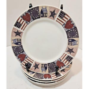 Può includere: Una pila di piatti in ceramica bianca con un bordo decorativo composto da cuori rossi, stelle blu, piccole case e motivi a bandiera americana. I piatti sono tenuti in posizione verticale da un supporto espositore in metallo nero.