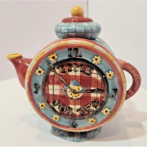May include: A ceramic teapot shaped like a round clock with a red and white checkered face. The clock face features black numbers, yellow flower accents, and the text TIME FOR TEA. It has a red spout, a red handle, and a blue and white lid.