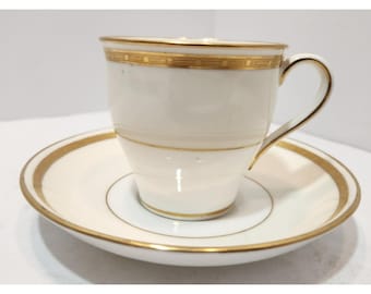 Puede incluir: Un juego de taza y platillo blancos con ribete dorado. La taza tiene un asa curvada y una banda dorada alrededor del borde. El platillo tiene un ribete dorado a juego. El juego es probablemente de porcelana o material similar.