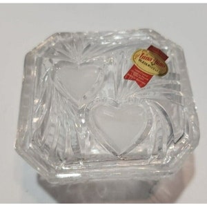 May include: A clear glass trinket box with a square shape and bevelled edges. The box has two heart-shaped indentations in the top and a label that reads "Anna Hutte Bleikristall." The label is red and white with gold lettering.