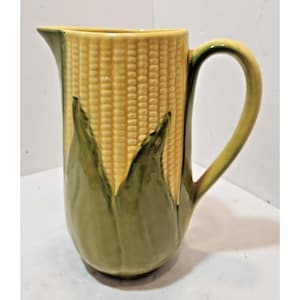 Puede incluir: Una jarra de cerámica con forma de mazorca de maíz, con un cuerpo amarillo texturizado de granos y hojas verdes que forman la base y el asa. La jarra presenta una forma cilíndrica alta con un pico vertedor en el borde y un asa curva.