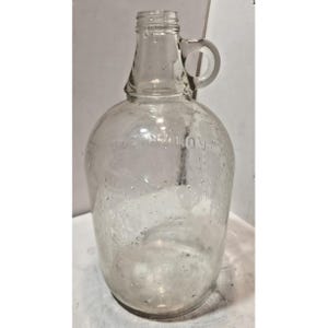 May include: A clear glass jug with a handle, embossed with the words "ONE GALLON". The jug has a rounded body and a narrow neck. The glass has small bubbles throughout.