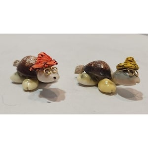 May include: Two small turtle figurines made from seashells. One turtle is wearing a red hat and glasses, the other is wearing a yellow hat and glasses.