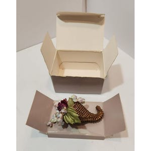 May include: A brown wicker cornucopia filled with purple, green, and blue fabric flowers. The cornucopia is in a light brown box with a white interior.