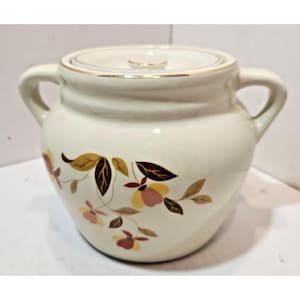 May include: A cream-colored ceramic sugar bowl with a matching lid and two side handles. The exterior features a decorative botanical pattern of stylized leaves and flowers in shades of brown, yellow, and muted pink, encircling the center.