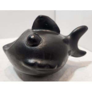 May include: A black ceramic fish figurine with a rounded body, a dorsal fin, and a tail fin. The fish has a large, round eye and a small mouth. The figurine is likely handmade and has a smooth, glossy finish.