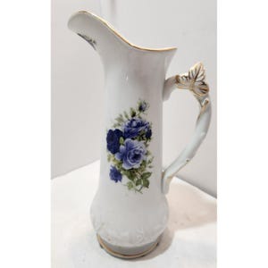 May include: White ceramic pitcher with a gold rim and handle, adorned with a cluster of blue roses and green leaves. The handle features a gold leaf design. A decorative floral pitcher for serving beverages.