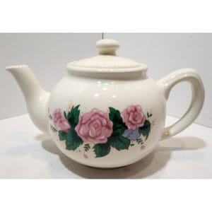 Puede incluir: Tetera de cerámica color crema con tapa y asa. La tetera está decorada con rosas rosas, hojas verdes y pequeñas flores azules. Tiene un pico para verter.