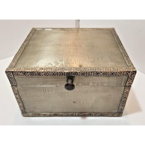 May include: A rectangular silver-toned box with a decorative border and a small, round knob. The box has a slightly textured surface and a hinged lid. The border features a repeating pattern of small, dark shapes. The box is closed.