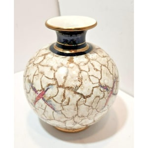 May include: A decorative ceramic vase with a round body and a narrow neck. The vase features a cracked, off-white surface with brown lines and accents of pink and blue. The neck is dark blue with gold trim.