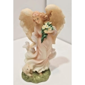 May include: A detailed figurine of an angel with peach-colored robes and large, textured wings. The angel holds a bouquet of white flowers with green stems. The base is a patch of green grass, and a small lamb is at the angel's feet.