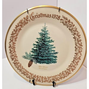 May include: A round ceramic plate with a cream-colored base and a gold-toned rim. The center features a green blue spruce tree with a brown pinecone. The text reads Christmas 1978, Blue Spruce, and Picea pungens in gold lettering.