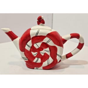 May include: A ceramic teapot shaped like a round peppermint sweet with a red and white spiral pattern. The teapot features a matching red and white striped handle and spout, with a small red and white knob on the lid. The finish is glossy.