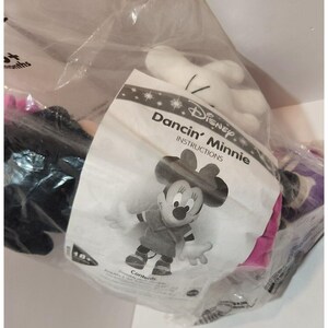May include: A plastic bag containing a Disney plush toy of Minnie Mouse. The toy is wearing a pink dress and has a black and white polka dot bow. The bag also contains instructions for the toy, which are titled "Dancin' Minnie Instructions".