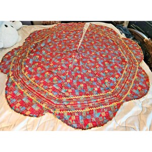 May include: A circular Christmas tree skirt featuring a vibrant patchwork pattern of red, blue, and green squares with festive motifs. The fabric is finished with a scalloped edge trimmed in gold rickrack, and it includes a slit for easy placement.