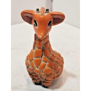 May include: A ceramic giraffe figurine with an orange and brown color scheme. The giraffe has large ears, black eyes, and a textured body. The figurine is a decorative item.