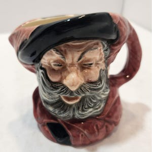 May include: A ceramic mug shaped like a bearded man's head, with a black hat and red robe. The mug has a handle and is painted with detailed facial features, including a long gray beard and closed eyes. The mug is likely a decorative item.