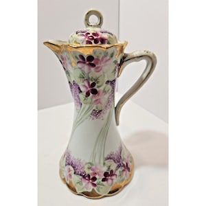May include: A porcelain pitcher with a lid, featuring a hand-painted floral pattern of purple and pink violets on a white background. The pitcher has a flared rim, a curved handle, and gold-painted trim along the edges and the lid handle.