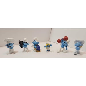 Puede incluir: Un conjunto de seis figuras azules de Pitufos, cada una con un sombrero blanco y un accesorio diferente. Los Pitufos sostienen un teléfono, una cesta de uvas, una mancuerna, un gorro de cocinero y un libro.