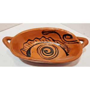 May include: An oval-shaped terracotta bowl with two side handles. The interior features a hand-painted dark brown spiral design and a jagged, wave-like pattern along the side. The clay has a natural reddish-brown finish with a smooth texture.