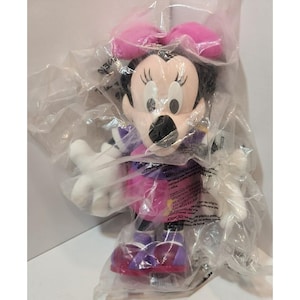 May include: A plush Minnie Mouse toy wearing a purple and pink dress with a white bow. The toy is packaged in clear plastic.