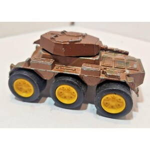 May include: A brown toy armored vehicle with six wheels and a rotating turret. The wheels have yellow centers and black tires. The vehicle has a weathered appearance, suggesting it is a vintage toy.