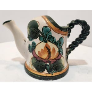 Puede incluir: Pequeña regadera decorativa de cerámica con boquilla blanca y asa trenzada. El cuerpo es de color crema con un diseño floral en naranja, rosa y verde. La base y el borde son de color verde oscuro.