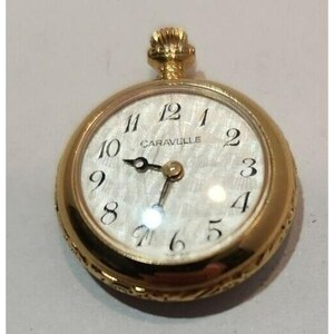 May include: A gold-toned pocket watch with a white face and black Roman numeral hour markers. The watch face has a textured, pearlescent finish and the words "Caravelle" printed on it.