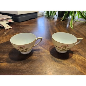 Könnte beinhalten: Zwei weiße Teetassen mit Blumendesigns und goldenen Verzierungen. Die Tassen stehen auf einer Holzoberfläche.