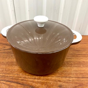 Puede incluir: Una olla de hierro fundido esmaltada marrón con un pomo de tapa blanco y asas blancas. La olla está sobre una superficie de madera.