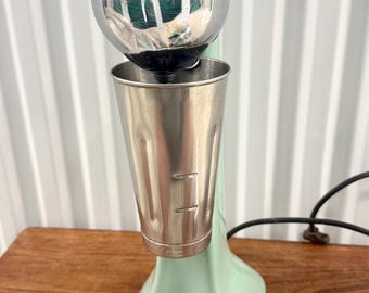Pode incluir: Uma misturadora de batidos Oster verde menta com uma caixa de motor em cromo polido com detalhes de linhas horizontais e um copo misturador de aço inoxidável removível fixado na frente. O aparelho tem uma base robusta e um cabo preto.