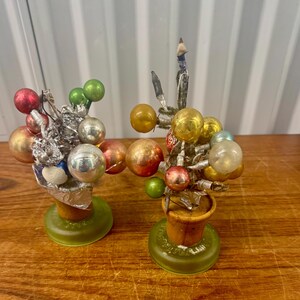 May include: Two decorative arrangements featuring clusters of small, round, multi-colored glass ornaments in shades of red, green, silver, and gold. Each arrangement is set in a tan, cone-shaped base mounted on a circular, translucent green stand.