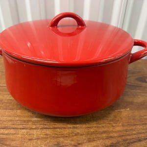 Könnte beinhalten: Ein leuchtend roter Dutch Oven mit passendem Deckel. Der Topf hat zwei seitliche Griffe und einen Schlaufengriff am Deckel. Der Topf steht auf einer Holzoberfläche.