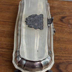 Puede incluir: Una caja rectangular plateada con una tapa con bisagras y una cresta decorativa en la parte superior. La caja tiene cuatro patas y un borde acanalado.