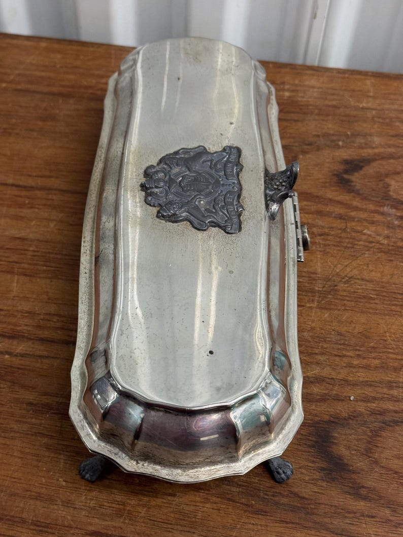 Puede incluir: Una caja rectangular plateada con una tapa con bisagras y una cresta decorativa en la parte superior. La caja tiene cuatro patas y un borde acanalado.