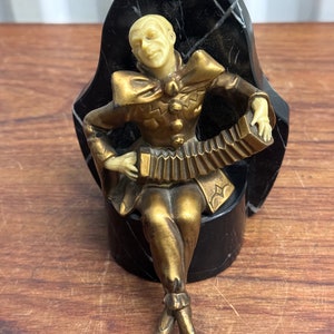 May include: A gold-coloured figurine of a jester playing an accordion, sitting on a black marble base. The jester is wearing a colourful costume with a large ruff collar.