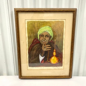 May include: A framed watercolor painting of a man wearing a green turban and a brown robe. He is sitting in a chair and holding a long pipe in his hand. The painting is titled "The Old Man with a Classic Eastern Pipe" and is signed by the artist "Dennis Yager 1963".