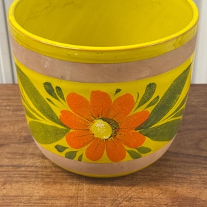 Könnte beinhalten: Handbemalter Keramik-Blumentopf. Der Topf ist überwiegend gelb mit einem beigen Band und einem großen orange-gelben Blumenmuster. Grüne Blätter umgeben die Blume. Der Topf steht auf einer Holzoberfläche.