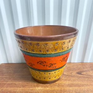 May include: A ceramic plant pot with a wide opening. It has a brown rim and a colourful design with orange, green, and yellow bands. The bands are decorated with floral and diamond patterns. The pot sits on a wooden surface.
