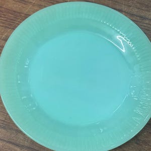 May include: A round, light mint green glass plate featuring a smooth, flat centre and a wide, textured rim with vertical ribbed detailing. The plate has a translucent, glossy finish and a slightly raised edge, showcasing a consistent colour throughout.