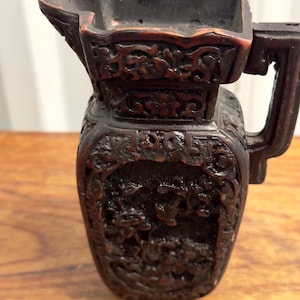 Könnte beinhalten: Ein dunkelbrauner Keramik-Krug mit aufwendigen Schnitzereien von Blumen und Ranken. Der Krug hat einen quadratischen Boden und einen breiten, abgerundeten Ausguss.