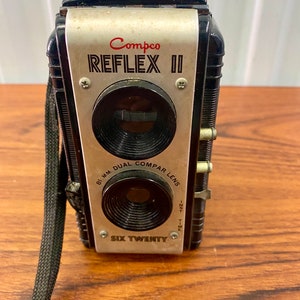 Puede incluir: Una cámara vintage Compco Reflex II negra y plateada con una lente comparadora doble. La cámara tiene una correa negra y dice "Six Twenty" en la parte delantera.