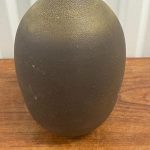 Könnte beinhalten: Eine dunkelbraune, tropfenförmige Keramikvase mit strukturierter Oberfläche. Die Vase hat einen Farbverlauf, der von einem helleren, goldbraunen Farbton oben zu einem dunkleren, fast schwarzbraunen Farbton am Boden übergeht. Die Vase steht auf einer Holzoberfläche.