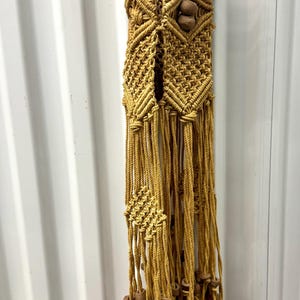 May include: A handcrafted macrame plant hanger in a warm, golden-yellow hue. The design features intricate knotting patterns, wooden beads, and a long fringe. This decorative piece is designed to hold a potted plant.
