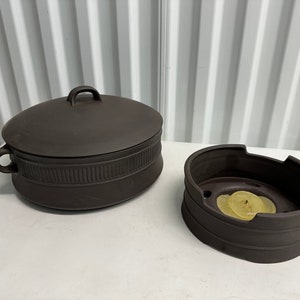 Può includere: Un piatto in ceramica marrone scuro con coperchio e una ciotola in ceramica marrone scuro abbinata con un bordo rialzato. La ciotola contiene una candela gialla.