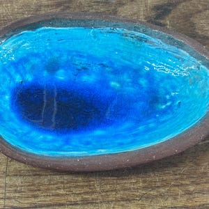 May include: Oval ceramic dish with a bright blue interior and a dark brown rim. The blue colour graduates from light to dark, creating depth. The dish is set against a wooden surface.
