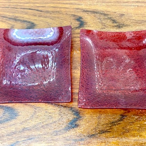 Könnte beinhalten: Zwei kleine, quadratische, rote Glasschalen mit einer strukturierten Oberfläche. Die Schalen sind leer und stehen auf einer Holzoberfläche.