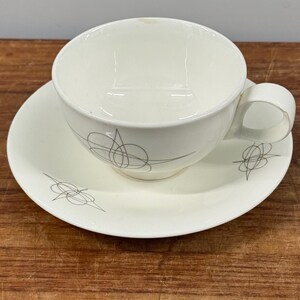 May include: Cream-colored teacup and saucer set. The cup has a rounded shape with a handle, and the saucer is circular. Both pieces are decorated with a delicate, gray abstract design. The set is made of ceramic.