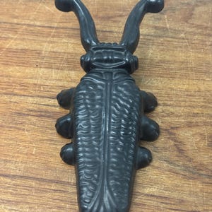 May include: A matte black cast iron beetle figurine featuring a textured, segmented shell and two prominent, curved antennae extending upward. The insect-shaped object has six legs and a detailed, ridged back, presenting a solid, heavy appearance.